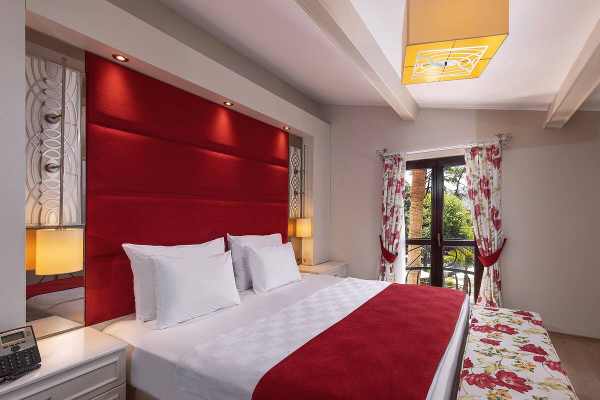 A hotel room with a red headboard, white bedding, a red throw blanket, and a floral-patterned bedspread.