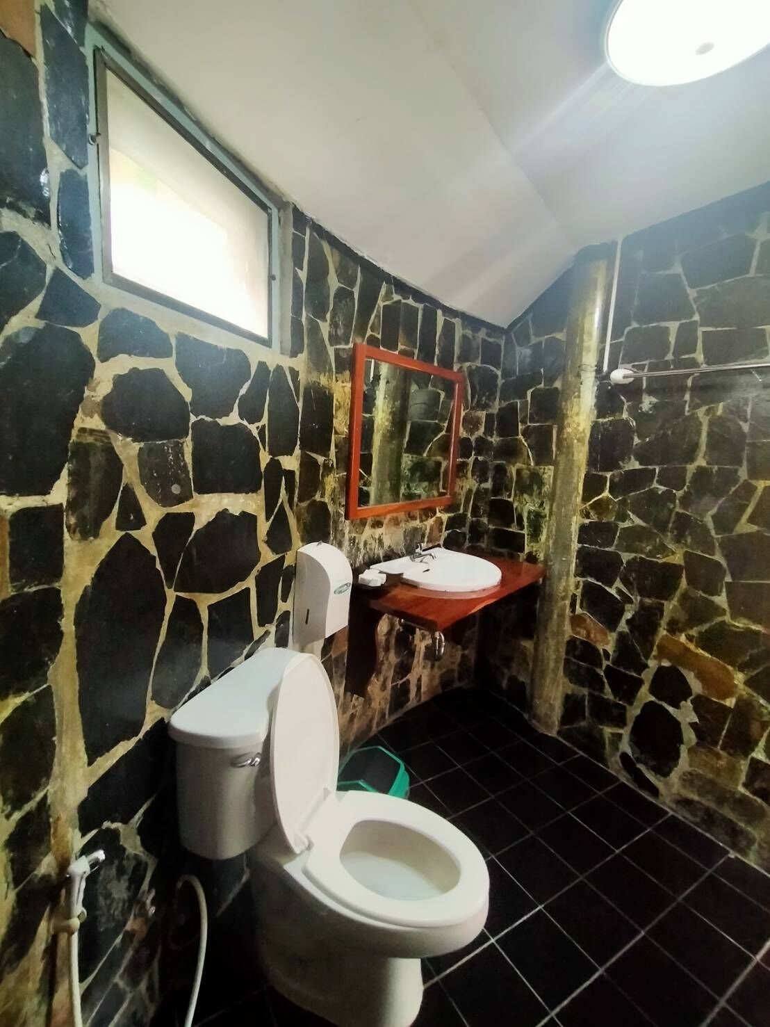 Un baño con pared de piedra, inodoro blanco, lavamanos con marco rojo y un espejo.