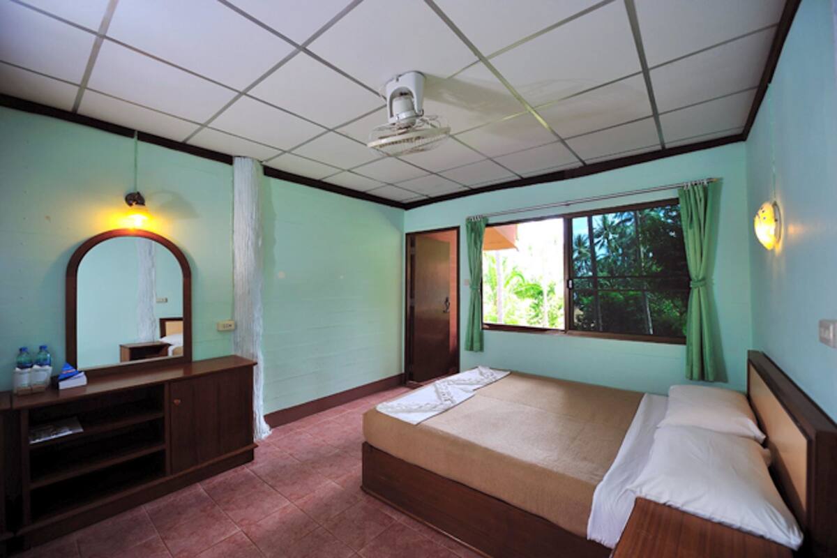 A bedroom with a bed, a bedside table, a mirror, a window with curtains, and a ceiling fan.
