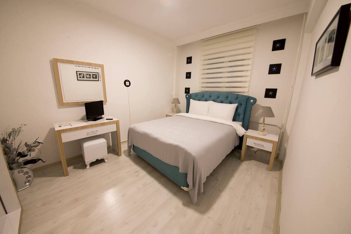 Ein Schlafzimmer mit Bett, Schreibtisch und Stuhl. Ein Fenster mit Jalousien, eine Lampe und ein Bilderrahmen sind an der Wand zu sehen.