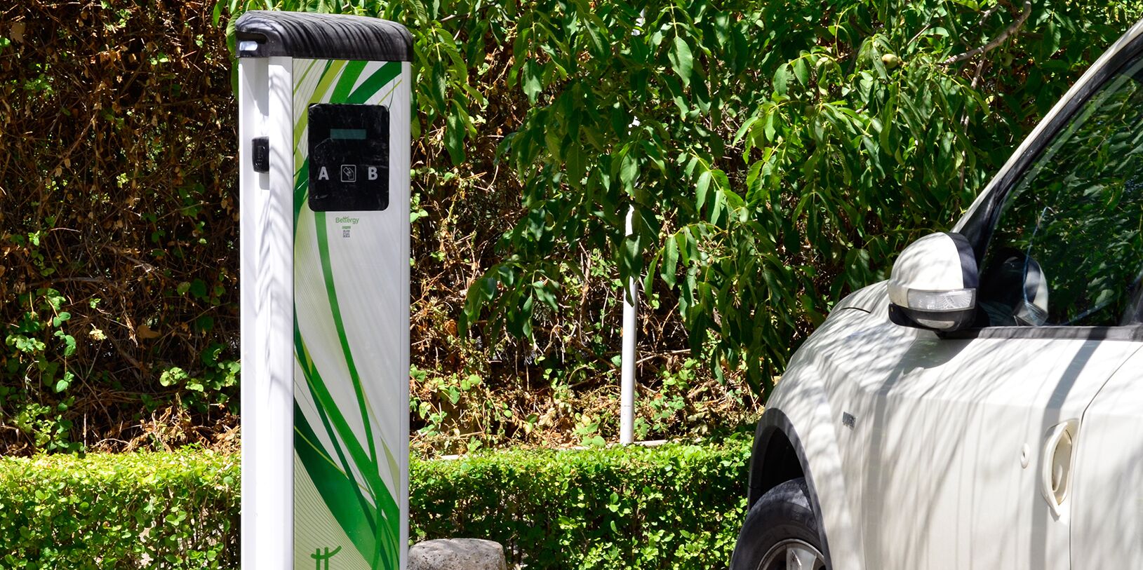 An electric vehicle charging station with a white and green design, located outdoors with greenery in the background.