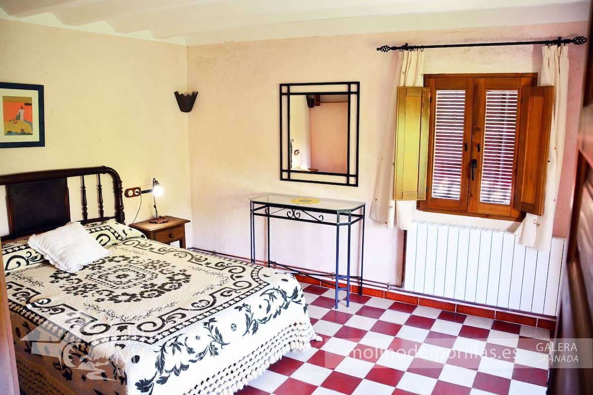 A bedroom with a checkered floor, a bed with a patterned bedspread, a bedside table with a lamp, a small table with a mirror, and a window with shutters.