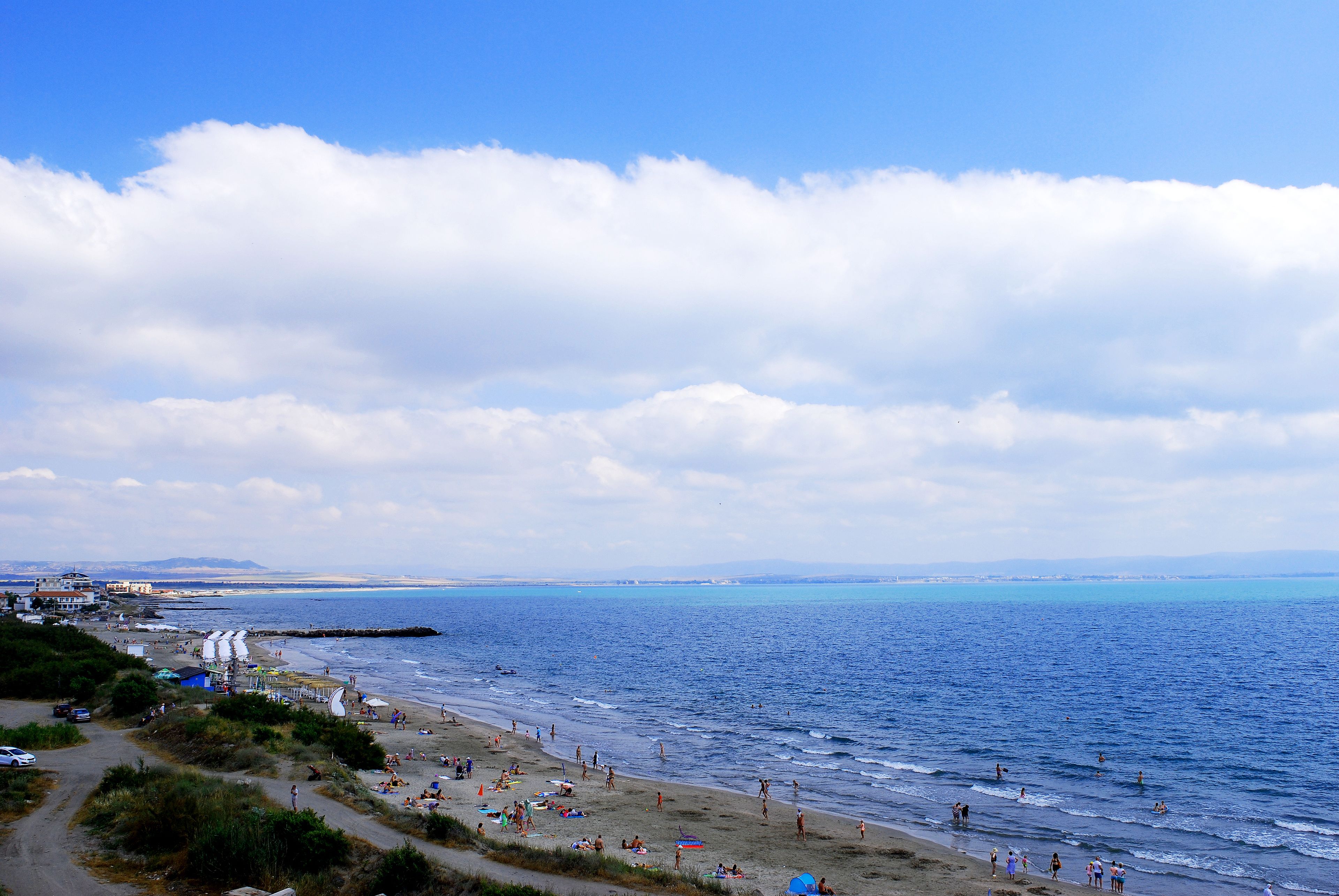 A coastal beach scene with a clear sky, sandy shore, and a few buildings in the background.