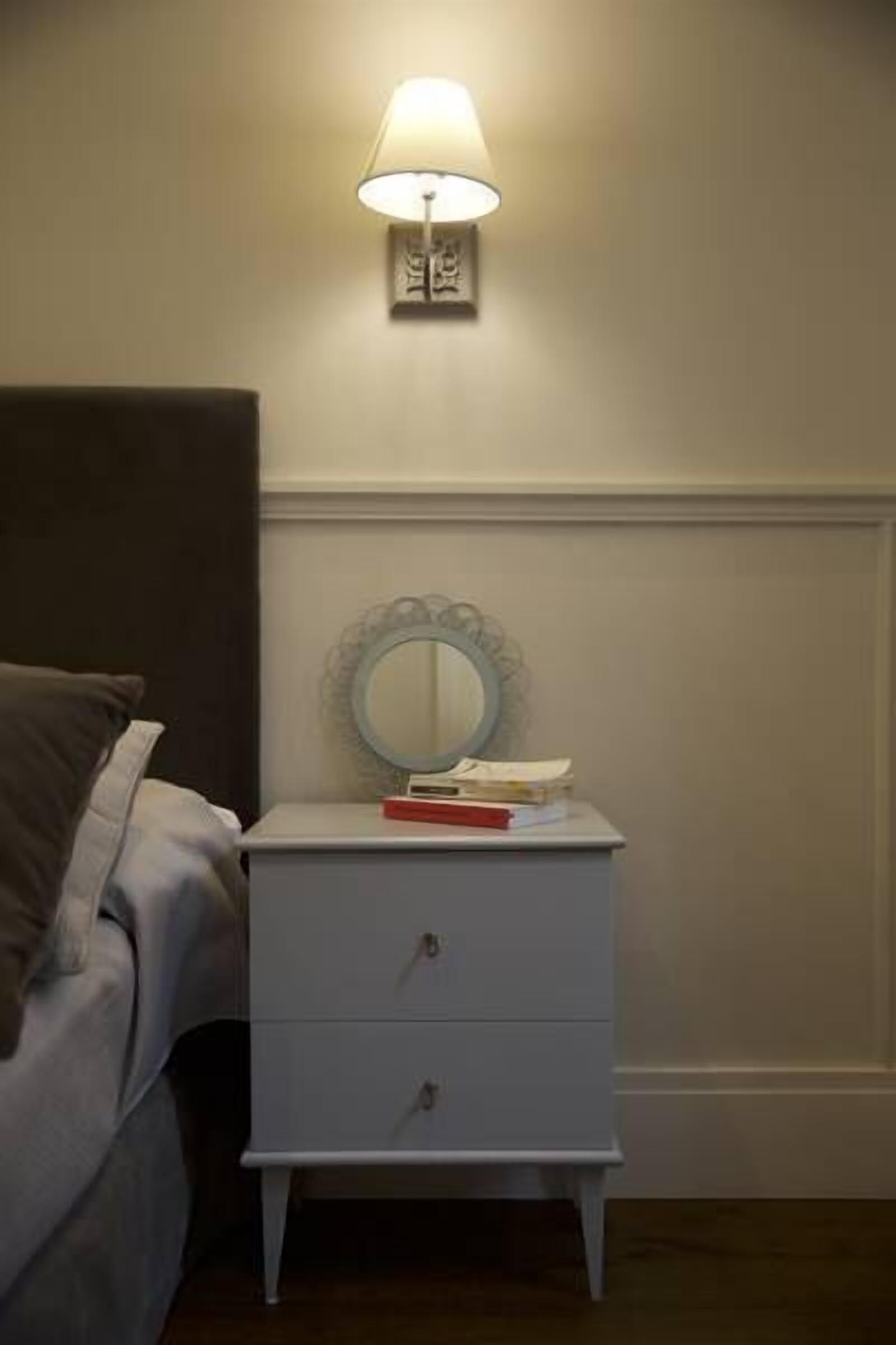 A bedroom with a bed, a bedside table with a lamp, a mirror, and a few books.