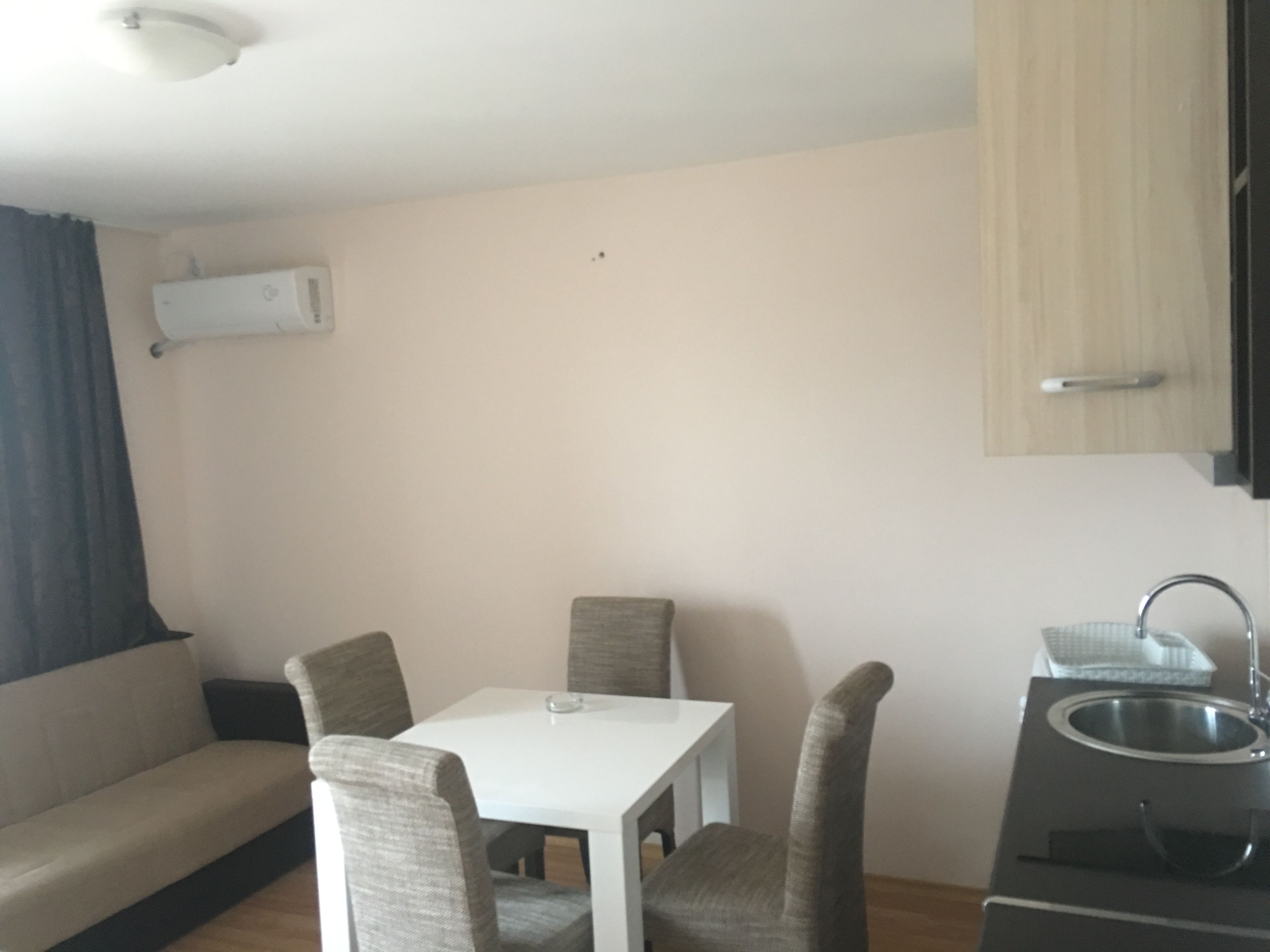 A small, modern kitchenette with a white countertop, a sink, and a stove. There is a white table with four chairs and a sofa with a dark throw pillow.