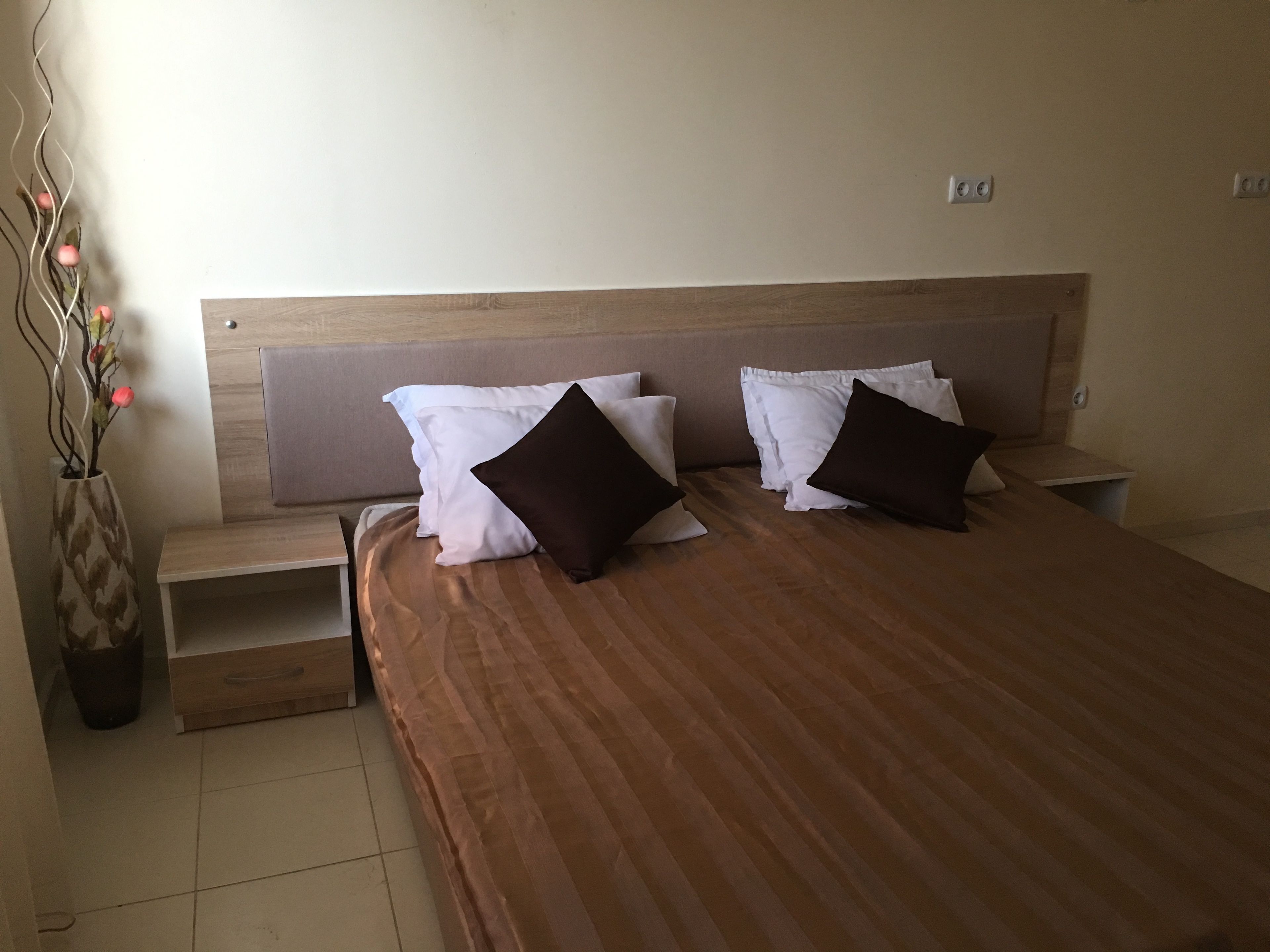 Een hotelkamer met een houten hoofdeinde, een bed met bruin beddengoed, witte en donkerbruine kussens, een nachtkastje en een vaas met gedroogde bloemen.