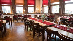 Un restaurant avec des tables et des chaises en bois, des chemins de table rouges et de grandes fenêtres ornées de rideaux rouges.