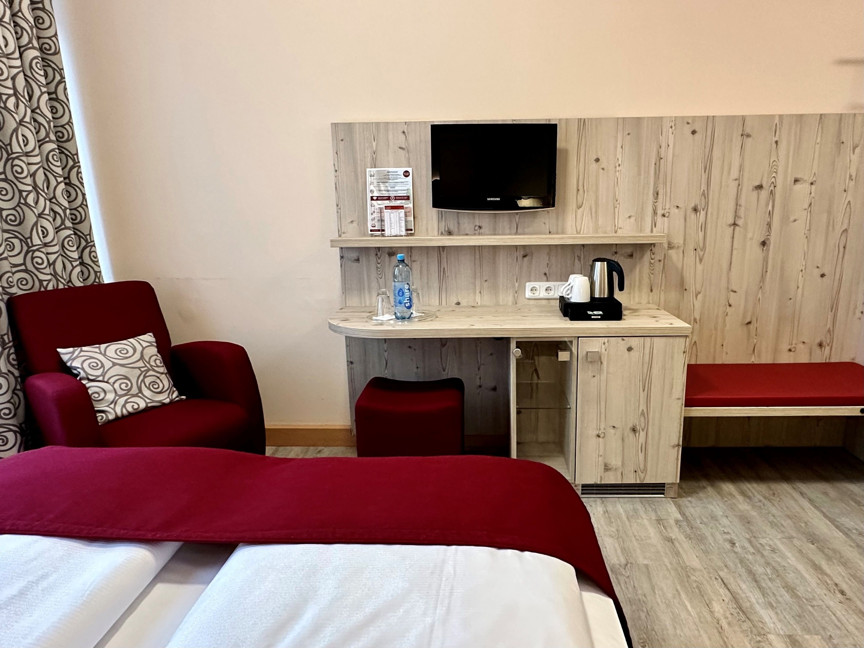 A hotel room with a bed, a desk with a television, a chair, a bench, and a water bottle.