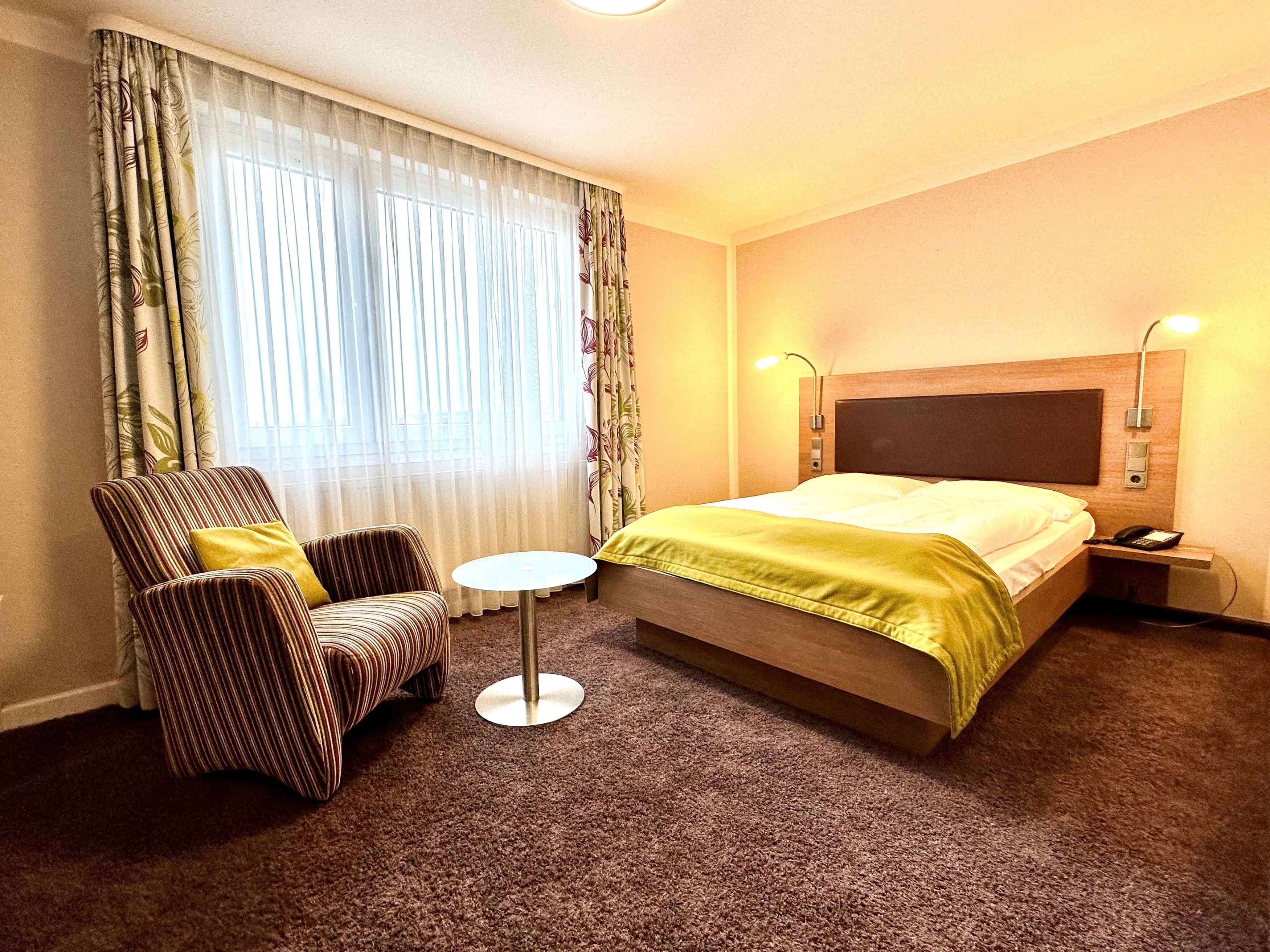 A hotel room with a bed, a chair, a small table, and curtains.