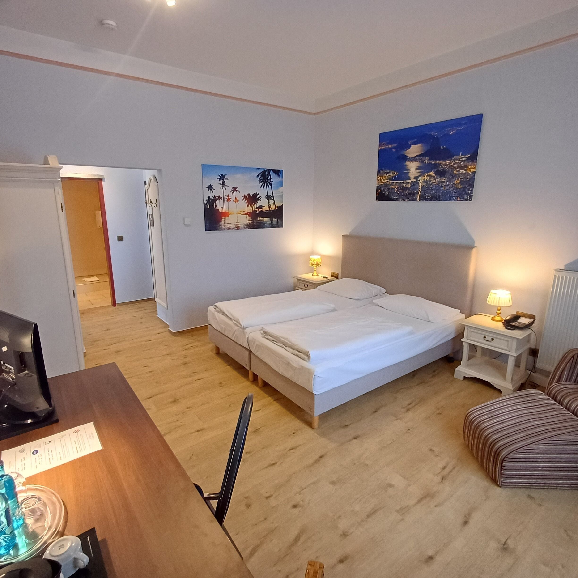 A hotel room with a bed, bedside tables, a desk with a computer, a chair, and two paintings on the walls.