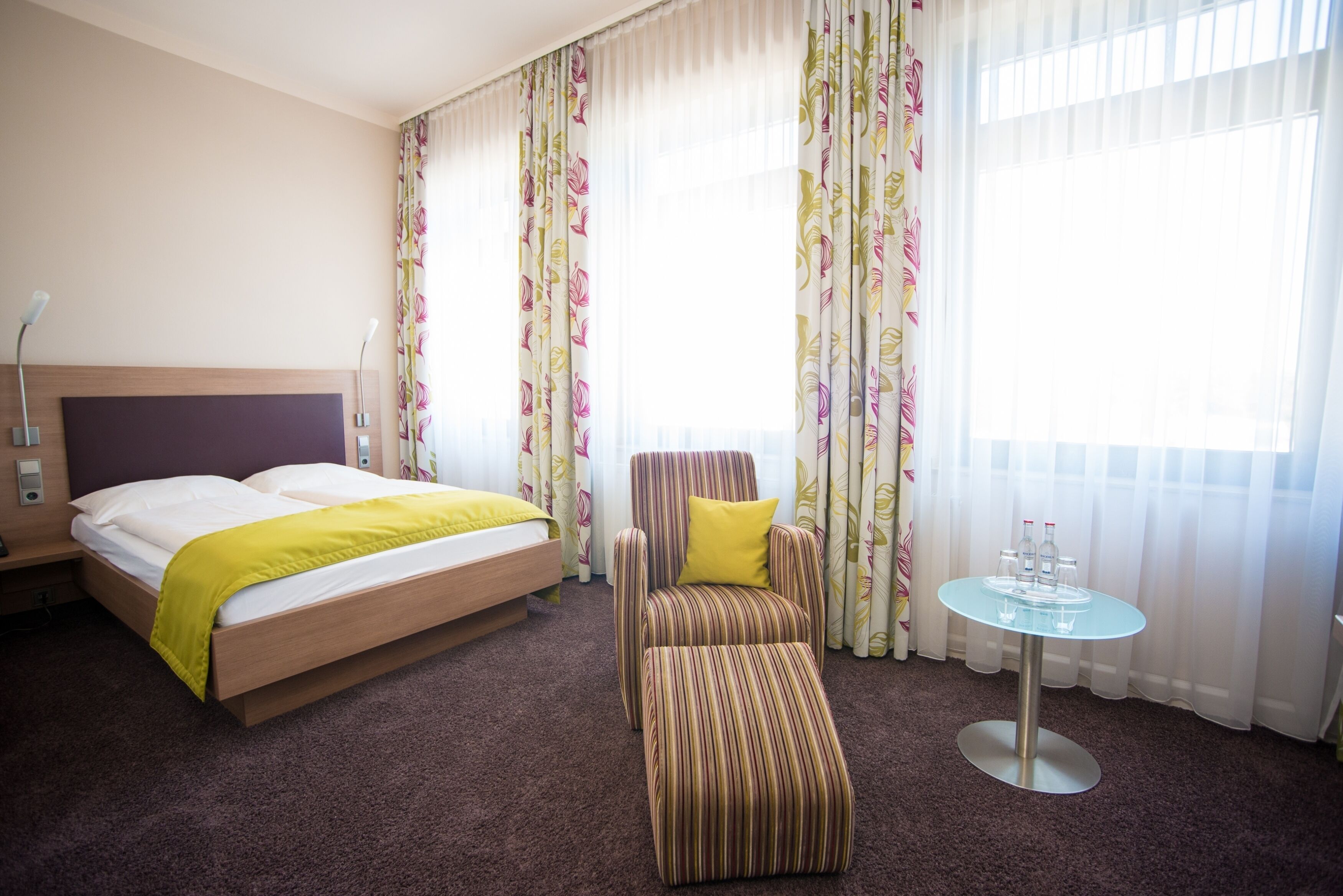 Ein Hotelzimmer mit Bett, einem gestreiften Sessel, einem kleinen Tisch mit Gläsern und Vorhängen mit Blumenmuster.