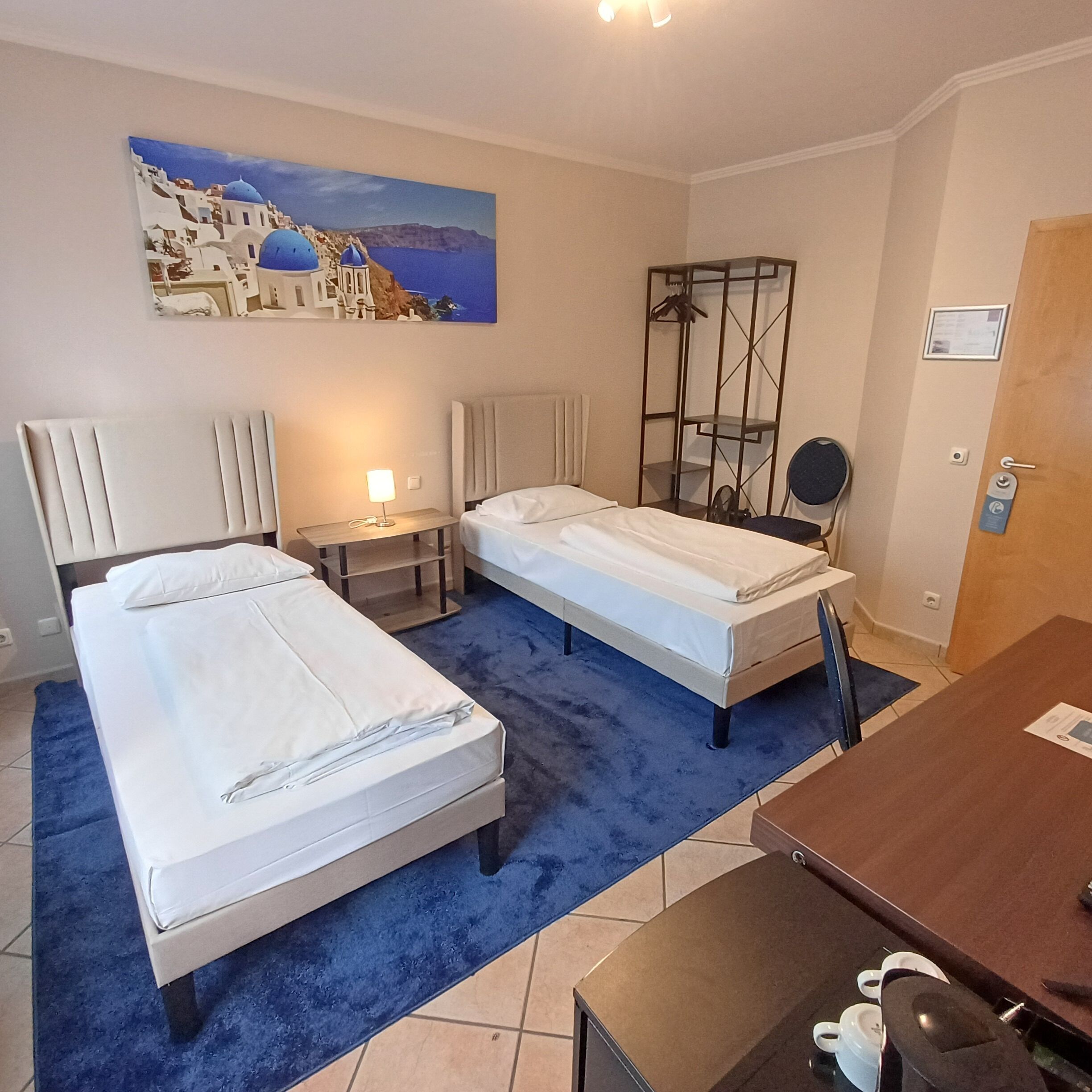 A hotel room with two beds, a desk, a chair, a small table, a painting of a coastal scene, and a wardrobe.
