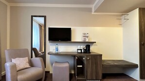 Une chambre d’hôtel compacte comprenant un canapé, une petite table avec une bouteille d’eau, une machine à café et un téléviseur à écran plat fixé au mur.
