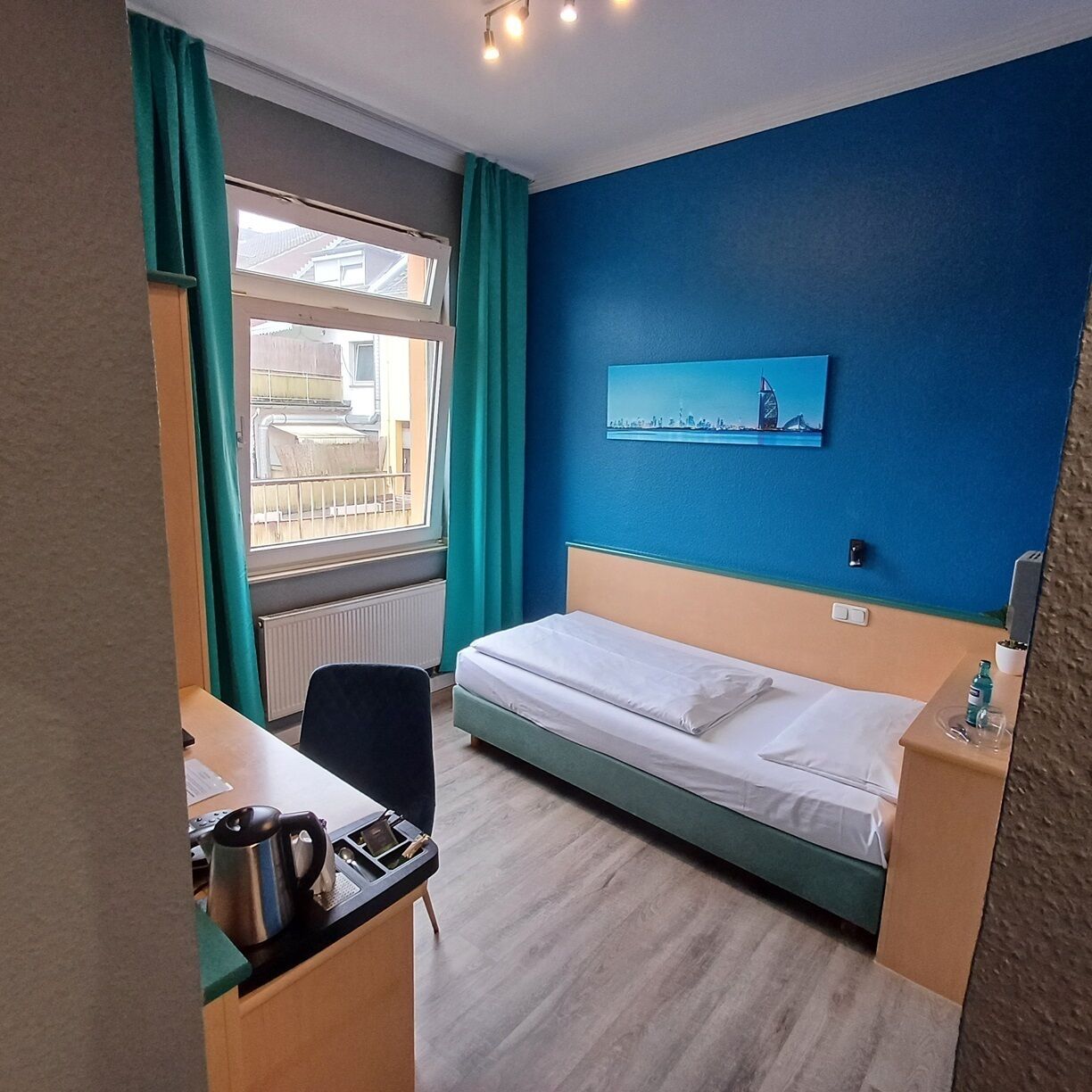 A hotel room with a bed, a desk with a kettle, a chair, and a window with curtains.