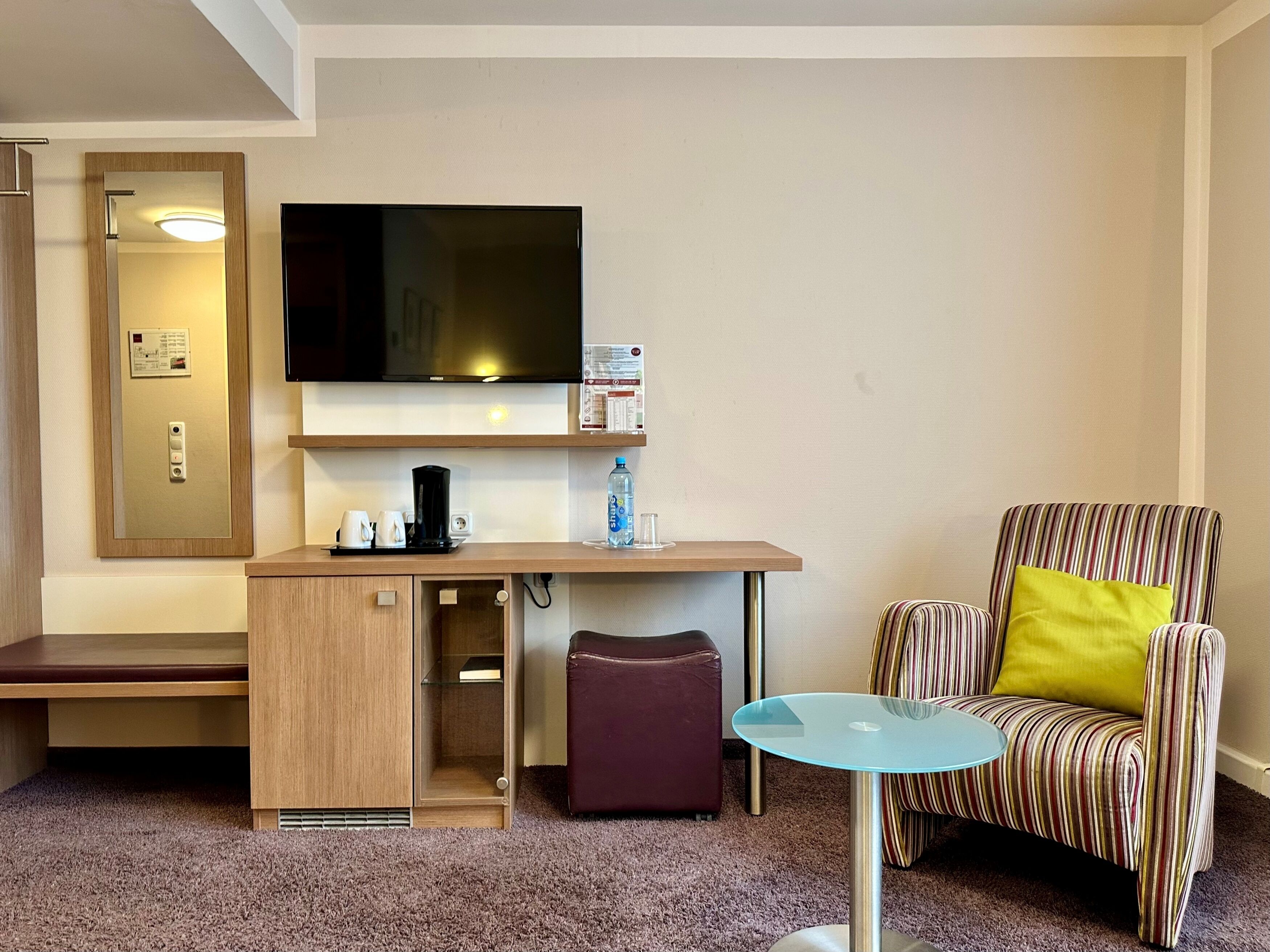 Ein kompaktes Hotelzimmer mit einem wandmontierten Flachbildfernseher, einem kleinen Holztisch mit Kaffeemaschine, einem Glastisch, einem gestreiften Sessel mit gelbem Kissen und einer Bank.