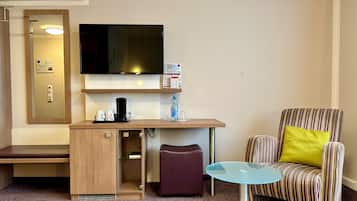 Ein kompaktes Hotelzimmer mit einem wandmontierten Flachbildfernseher, einem kleinen Holztisch mit Kaffeemaschine, einem Glastisch, einem gestreiften Sessel mit gelbem Kissen und einer Bank.