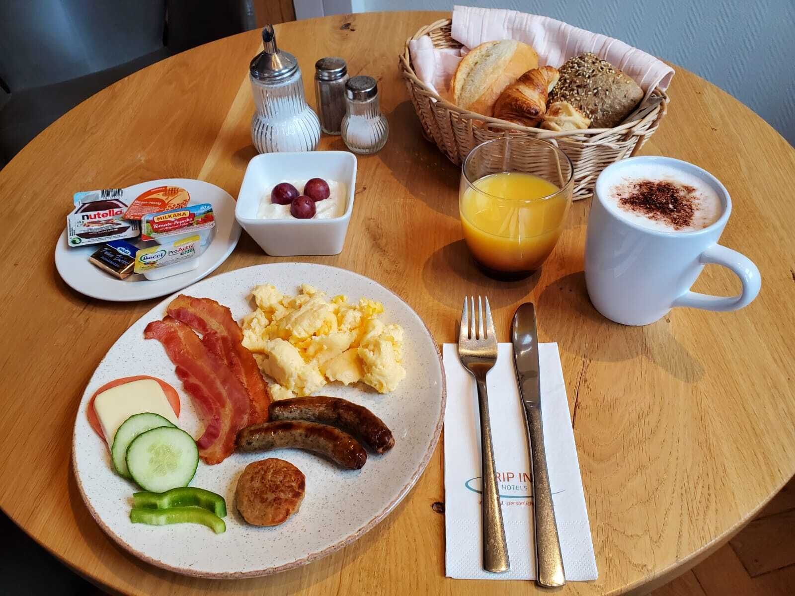 Una mesa de desayuno con huevos revueltos, tocino, salchichas y verduras. También hay condimentos, pan y una taza de café.