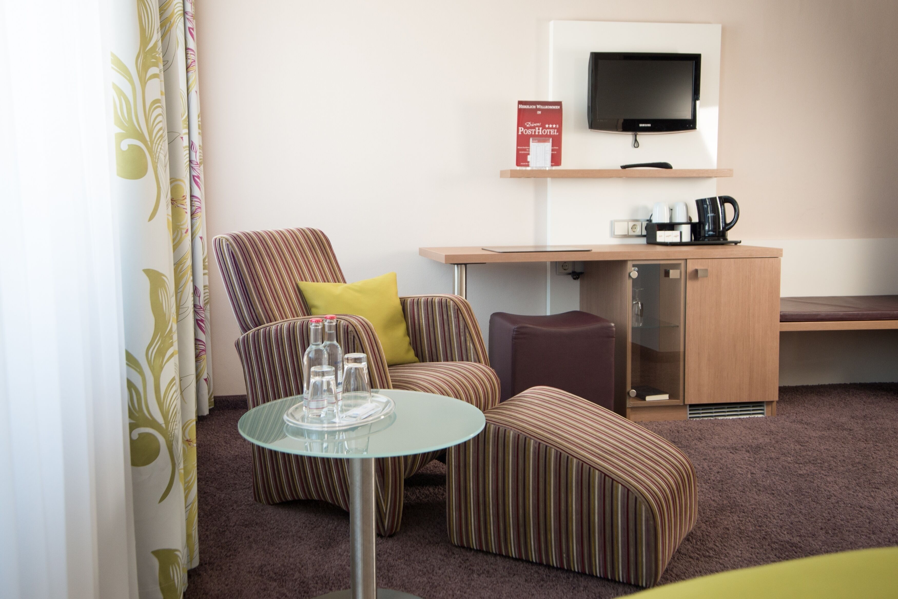 A hotel room with a striped armchair, a small round table with glasses, a television, a coffee maker, and a cabinet.