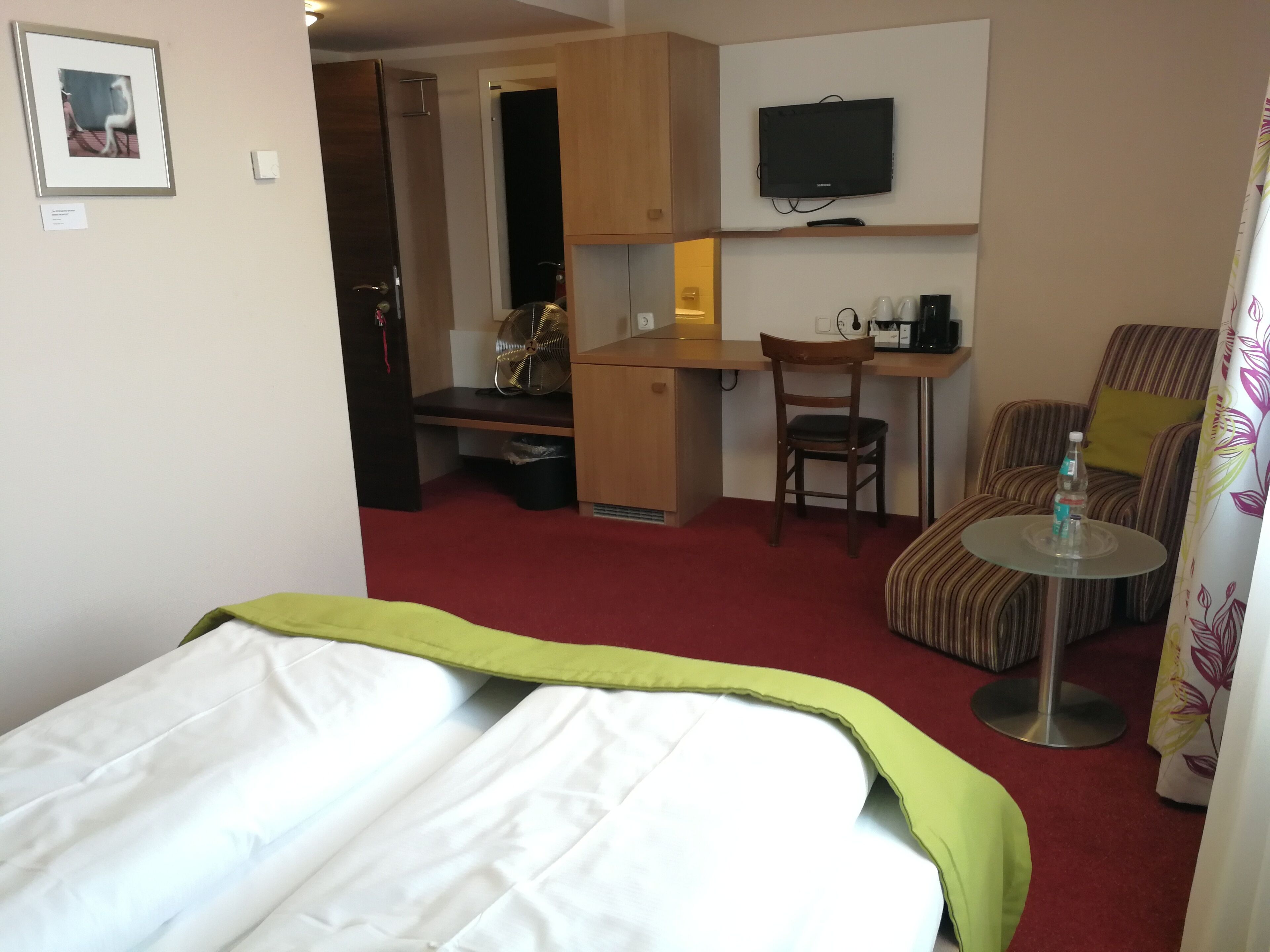 A hotel room with a bed, a desk, a chair, a TV, and a small table with a bottle.