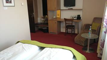 A hotel room with a bed, a desk, a chair, a TV, and a small table with a bottle.