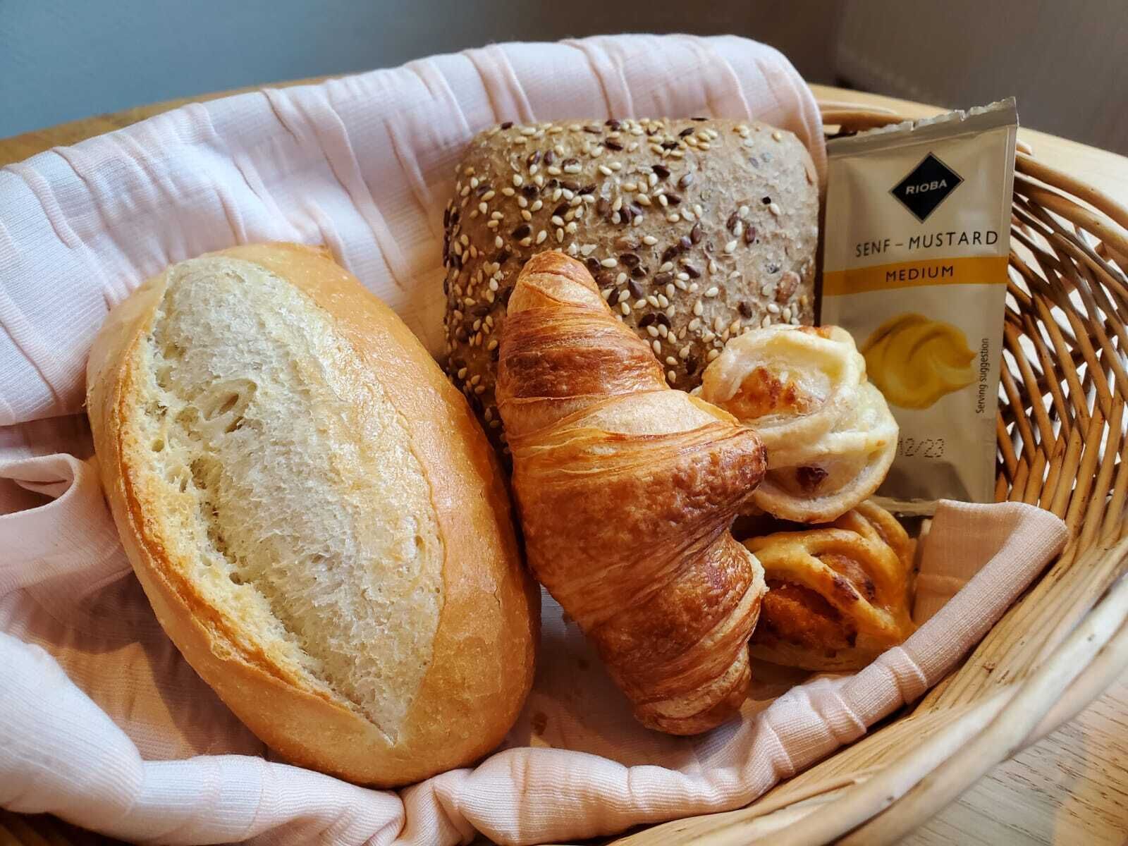 A basket containing a baguette, a croissant, a loaf of multigrain bread, and a packet of Senf-Mustard.