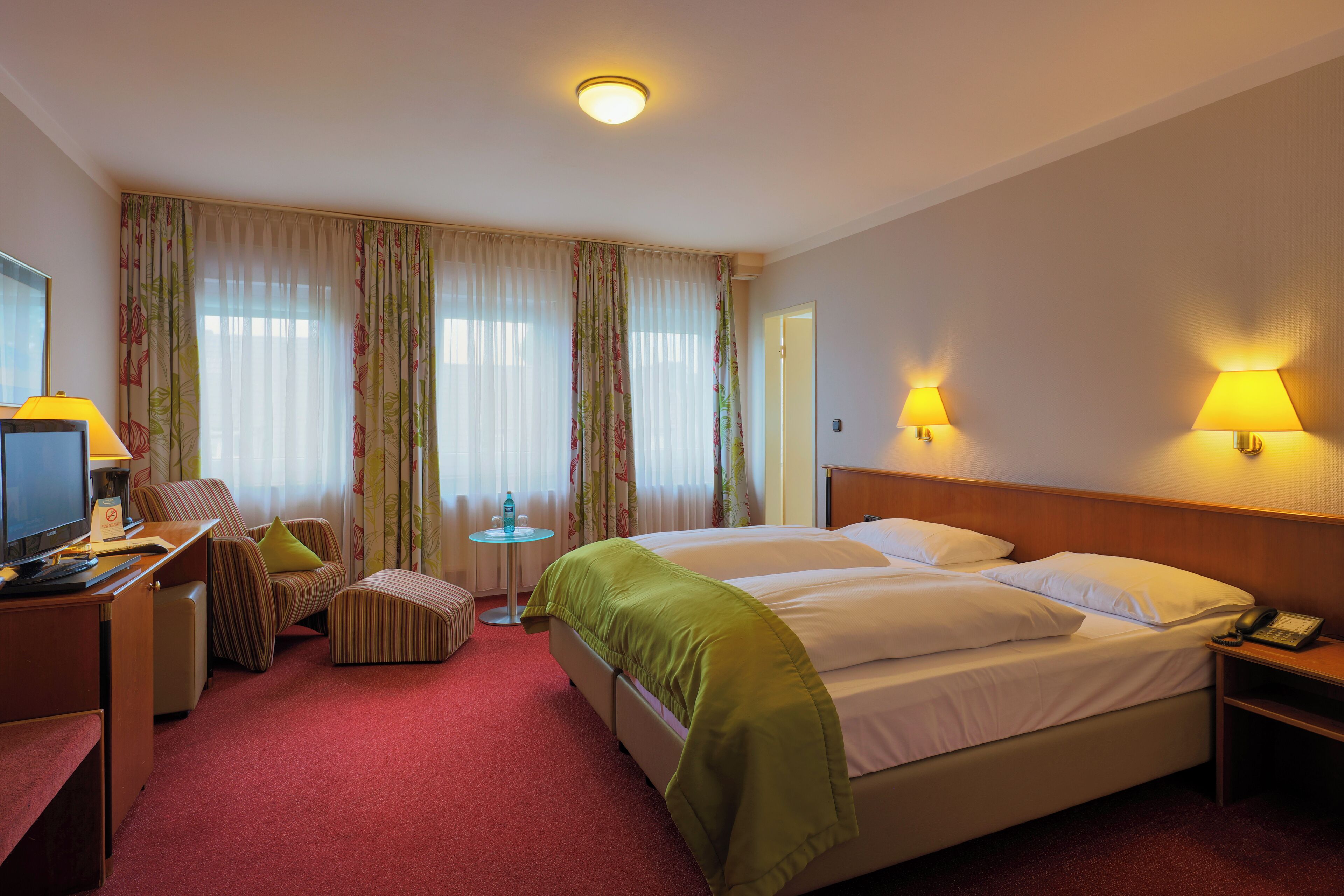 A hotel room with a bed, a desk with a computer, a chair, a small table with a bottle, and curtains.