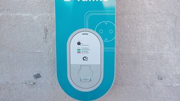 Una estación de carga para vehículos eléctricos de color verde azulado, con pantalla digital y enchufe de carga, montada en una pared de concreto.