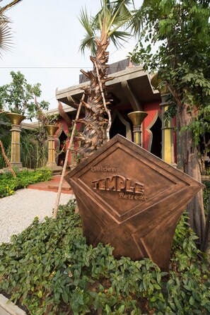 Un gran letrero metálico rústico con la inscripción “Temple Retreat” grabado, rodeado de exuberante follaje y un edificio tradicional al fondo.