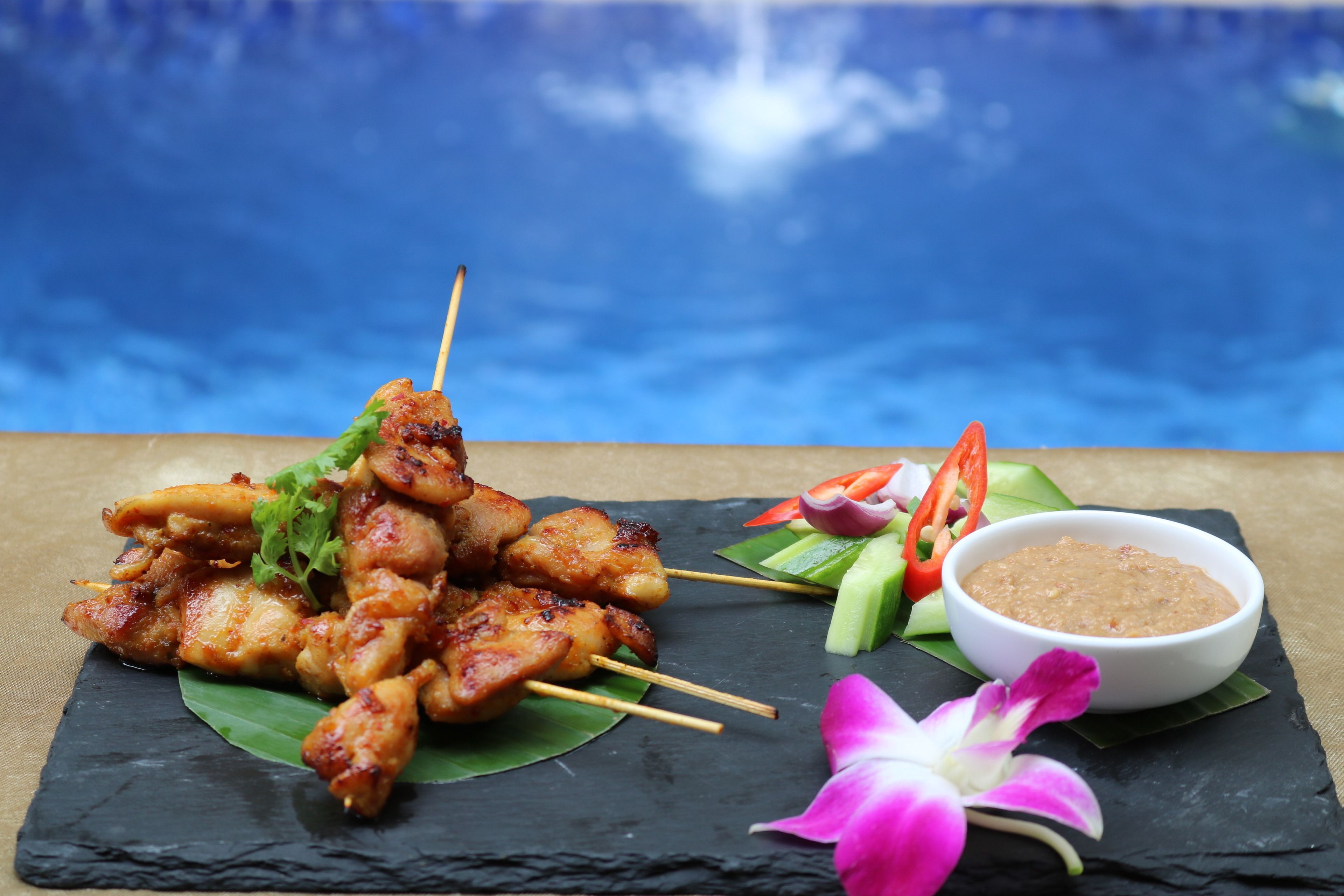Une assiette de brochettes de poulet grillées, accompagnées d’une sauce pour tremper et de quelques garnitures, le tout présenté avec une vue sur la piscine en arrière-plan.