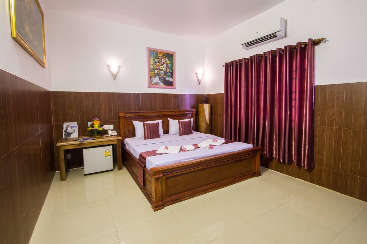 A hotel room with a wooden bed, a small desk with a refrigerator, a painting on the wall, and a window with curtains.