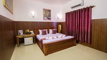 A hotel room with a wooden bed, a small desk with a refrigerator, a painting on the wall, and a window with curtains.