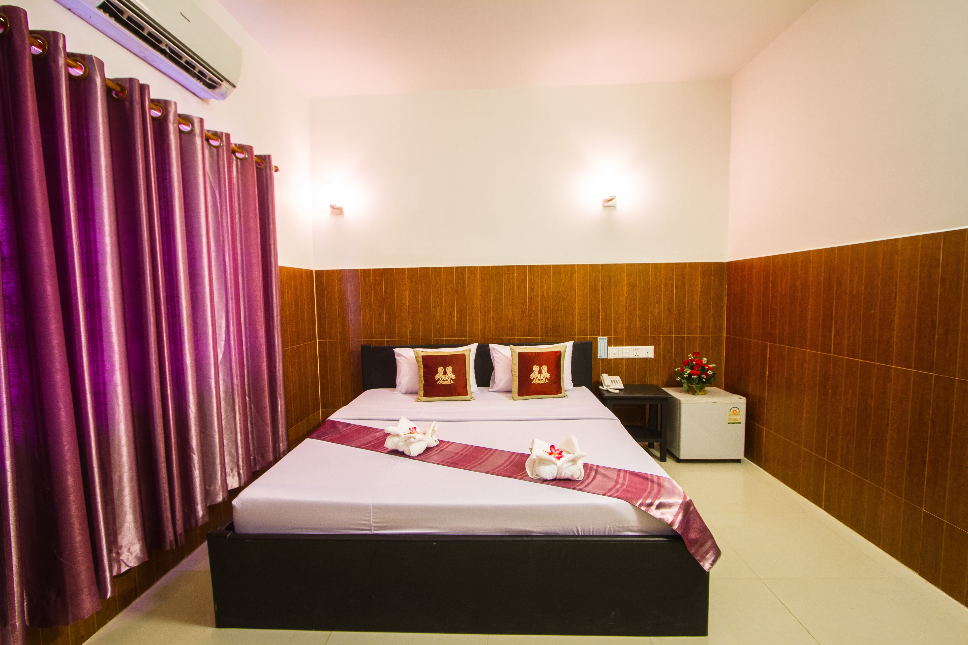 Ein Hotelzimmer mit einem großen Bett, Holzvertäfelung an den Wänden, einem Nachttisch mit Telefon und einem Blumenstrauß, sowie violetten Vorhängen.