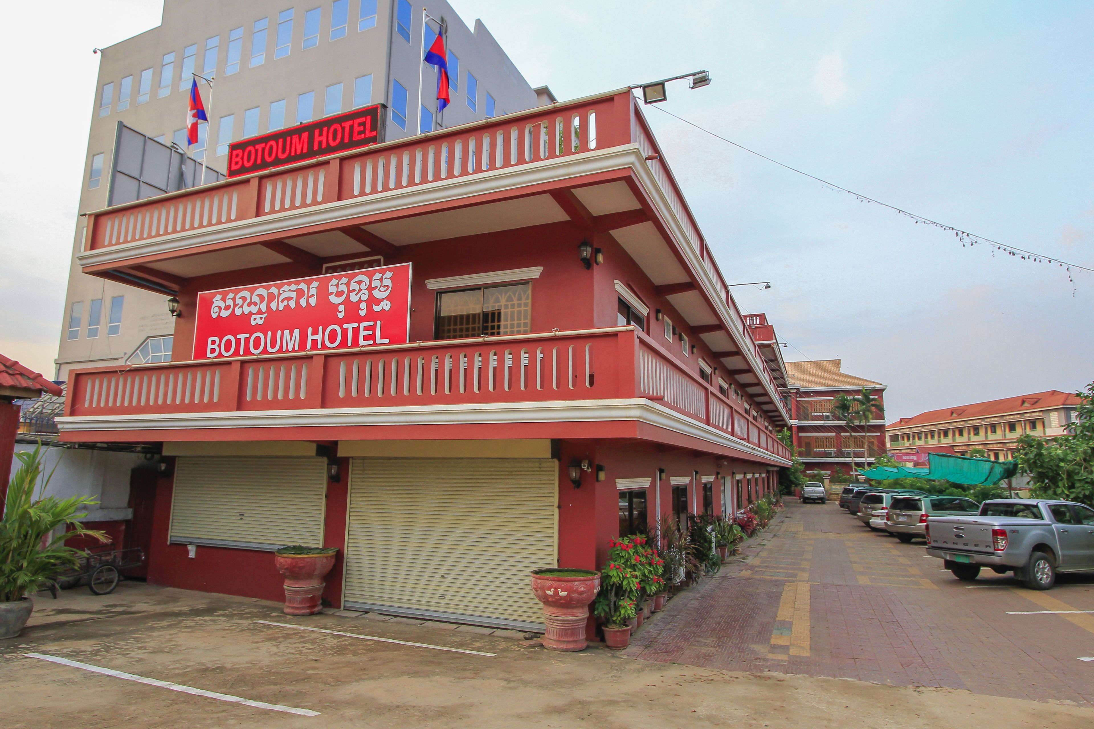 A two-story BOTOUM HOTEL with a red facade, multiple balconies, and a parking area with cars.