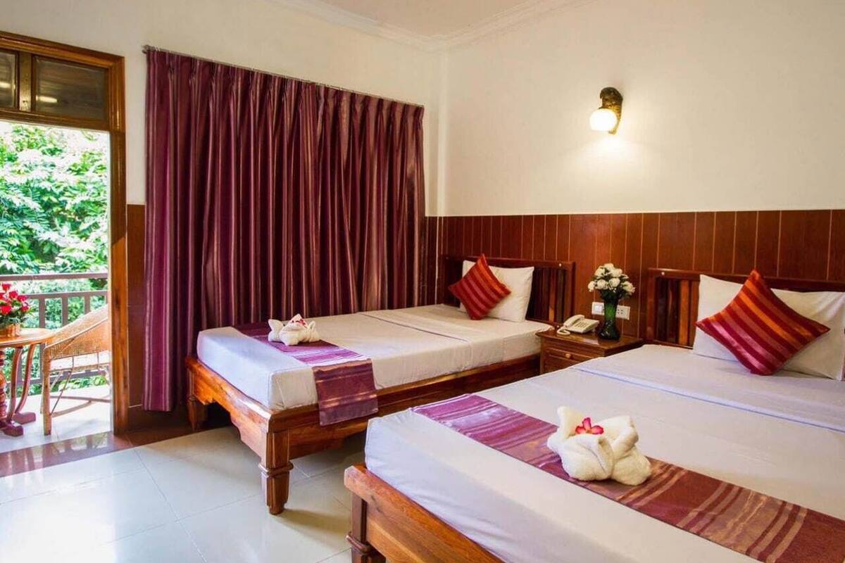 A hotel room with two beds, a wooden headboard, a small table with a chair, a window with curtains, and a balcony.