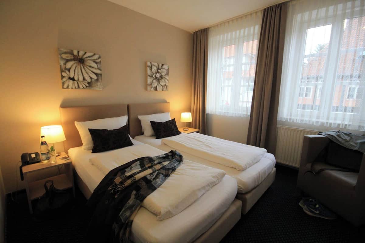 Habitación de hotel con dos camas, un escritorio con lámpara, una silla y obras de arte en las paredes.