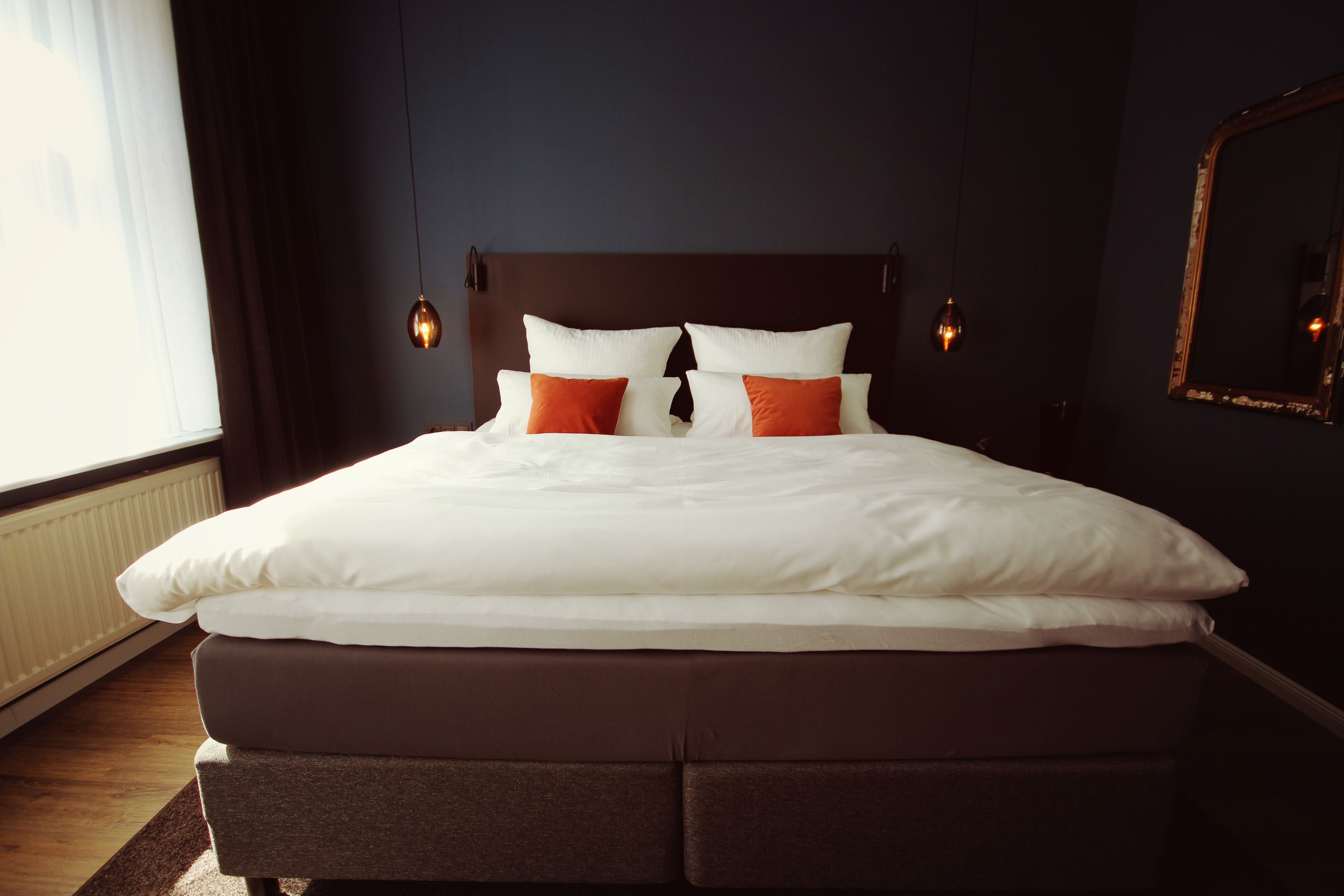 Een opgemaakt bed met witte en oranje kussens, een donker hoofdbord en wandlampen.