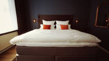 Een opgemaakt bed met witte en oranje kussens, een donker hoofdbord en wandlampen.