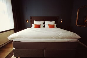 Una cama bien hecha con almohadas blancas y anaranjadas, un cabecero oscuro y luces fijadas a la pared.
