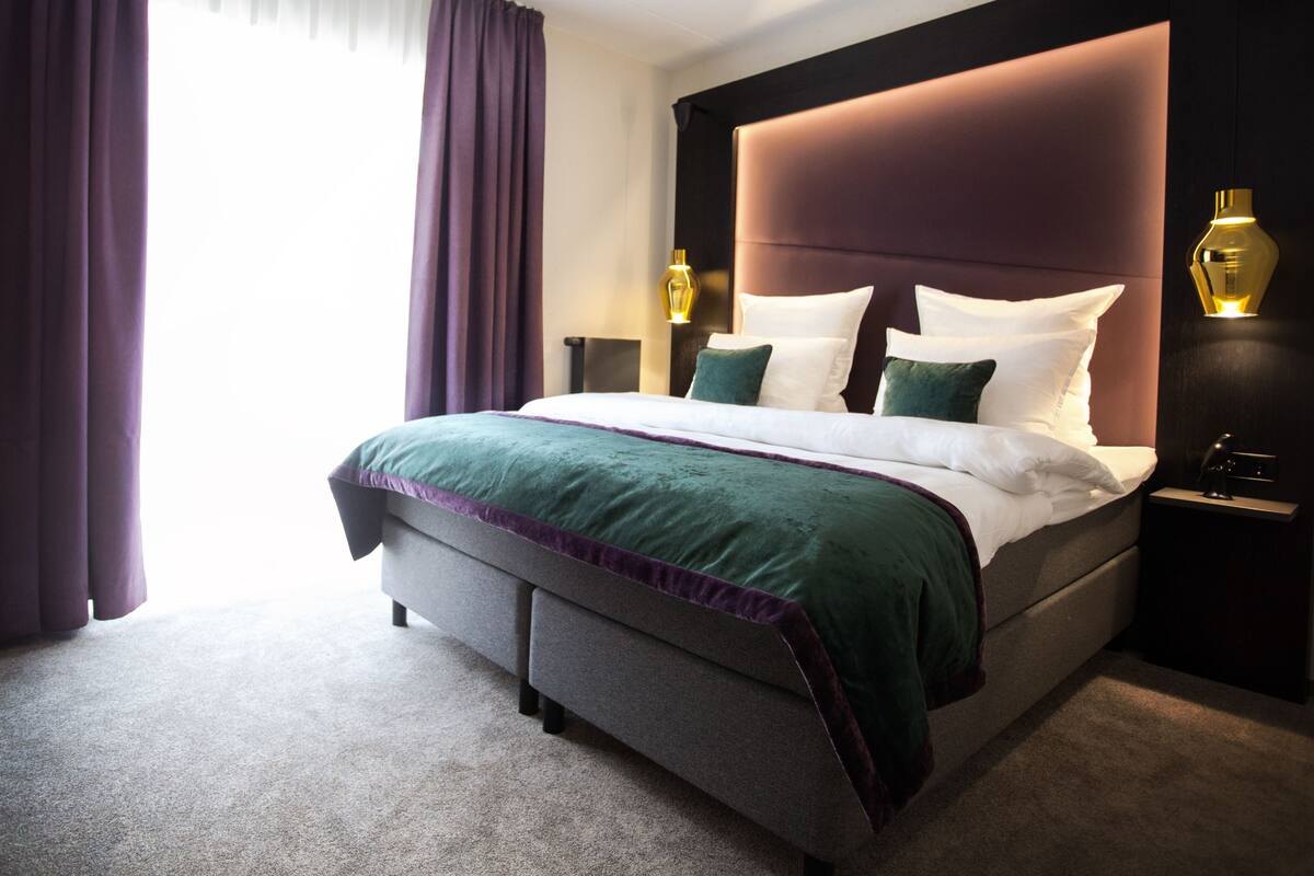 Una habitación de hotel con una cama grande, cabecera, dos lámparas de noche y mesitas de noche.