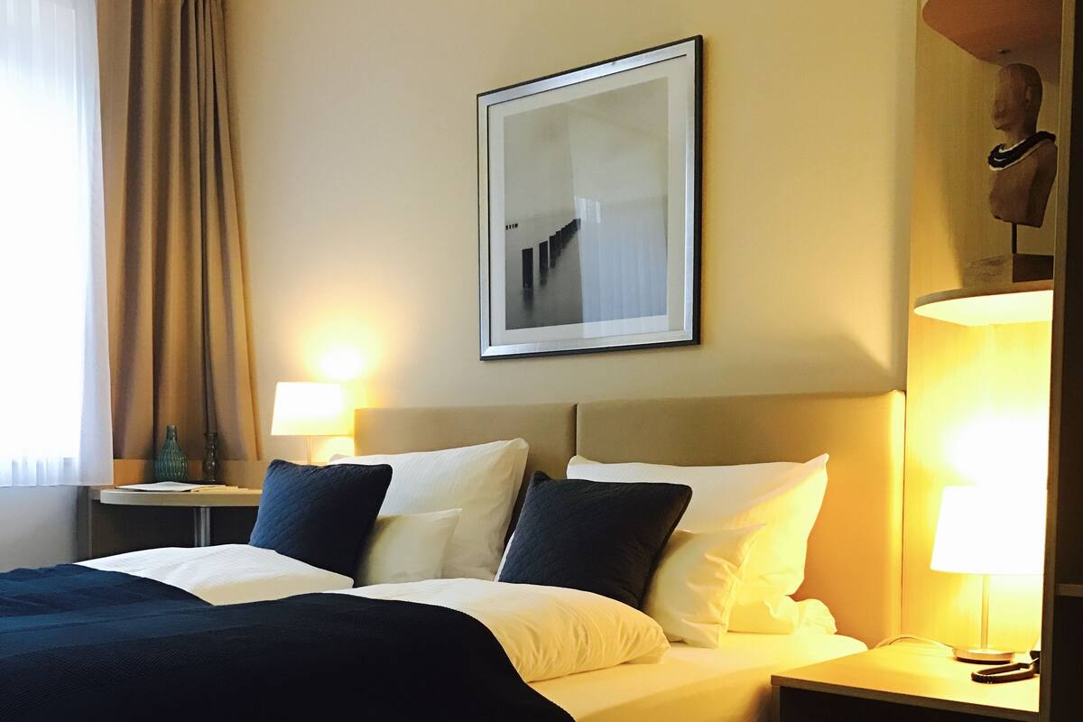 Habitación de hotel con cama, dos mesitas de noche, lámpara, un cuadro en la pared y ventana con cortinas.
