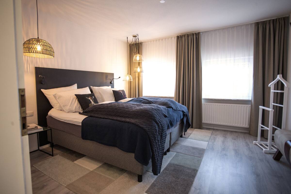 Una habitación de hotel moderna con una cama grande, una mesita de noche, un armario y una ventana con cortinas.