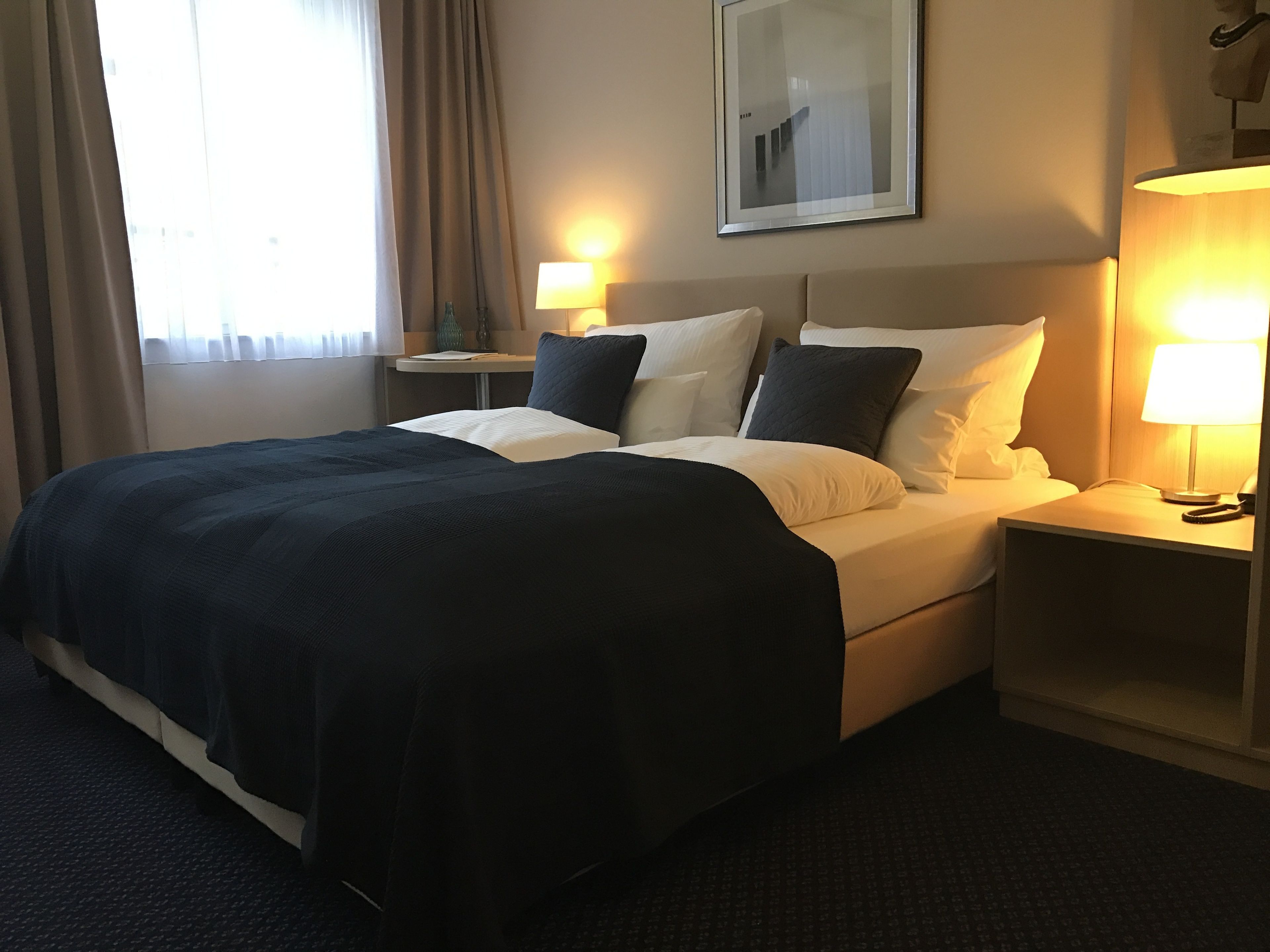 Ein Hotelzimmer mit einem großen Bett, zwei Nachttischen mit Lampen, einem Spiegel an der Wand und einem Fenster mit Vorhängen.