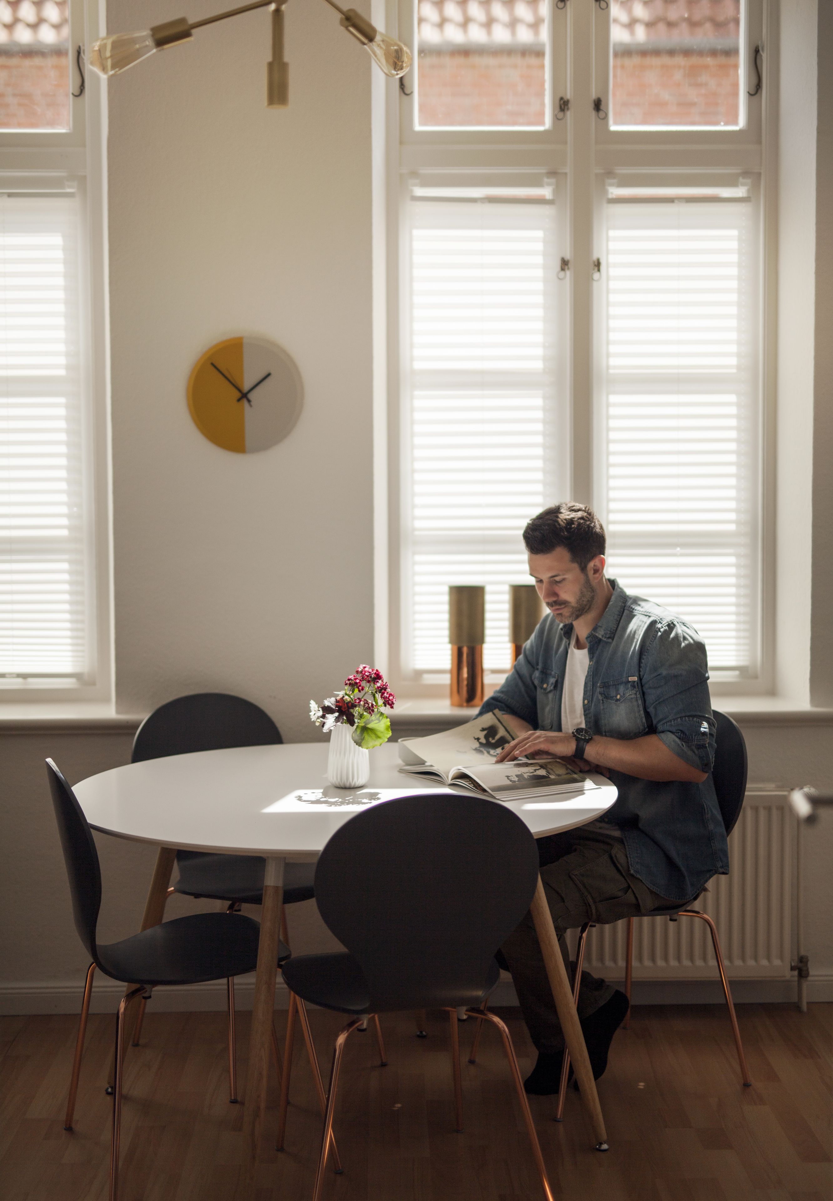 Een man zit aan een ronde eettafel met een wit tafelkleed, een boek lezend. Op de tafel staat een vaas met bloemen, en erboven een wandklok. De kamer heeft grote ramen met zonwering, en er is een radiator aan de muur te zien.