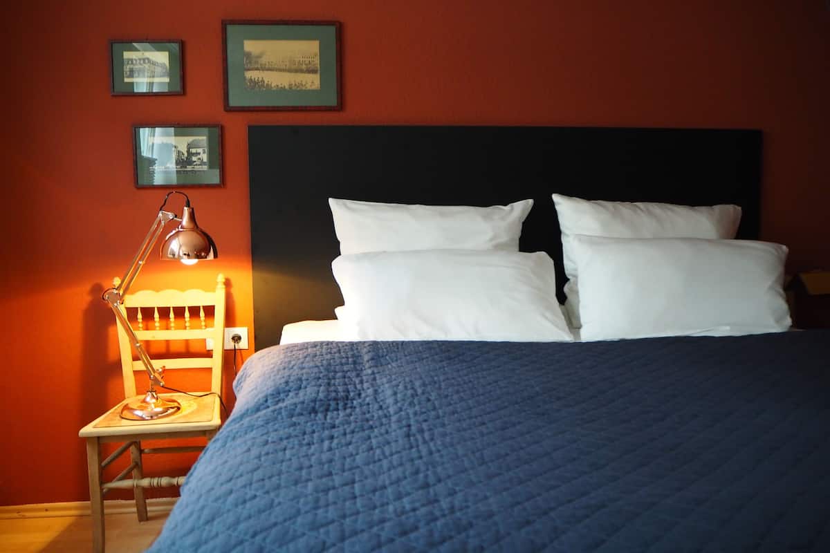 Una cama bien tendida con almohadas blancas y colcha azul, una pequeña silla de madera con una lámpara, y dos cuadros en la pared.