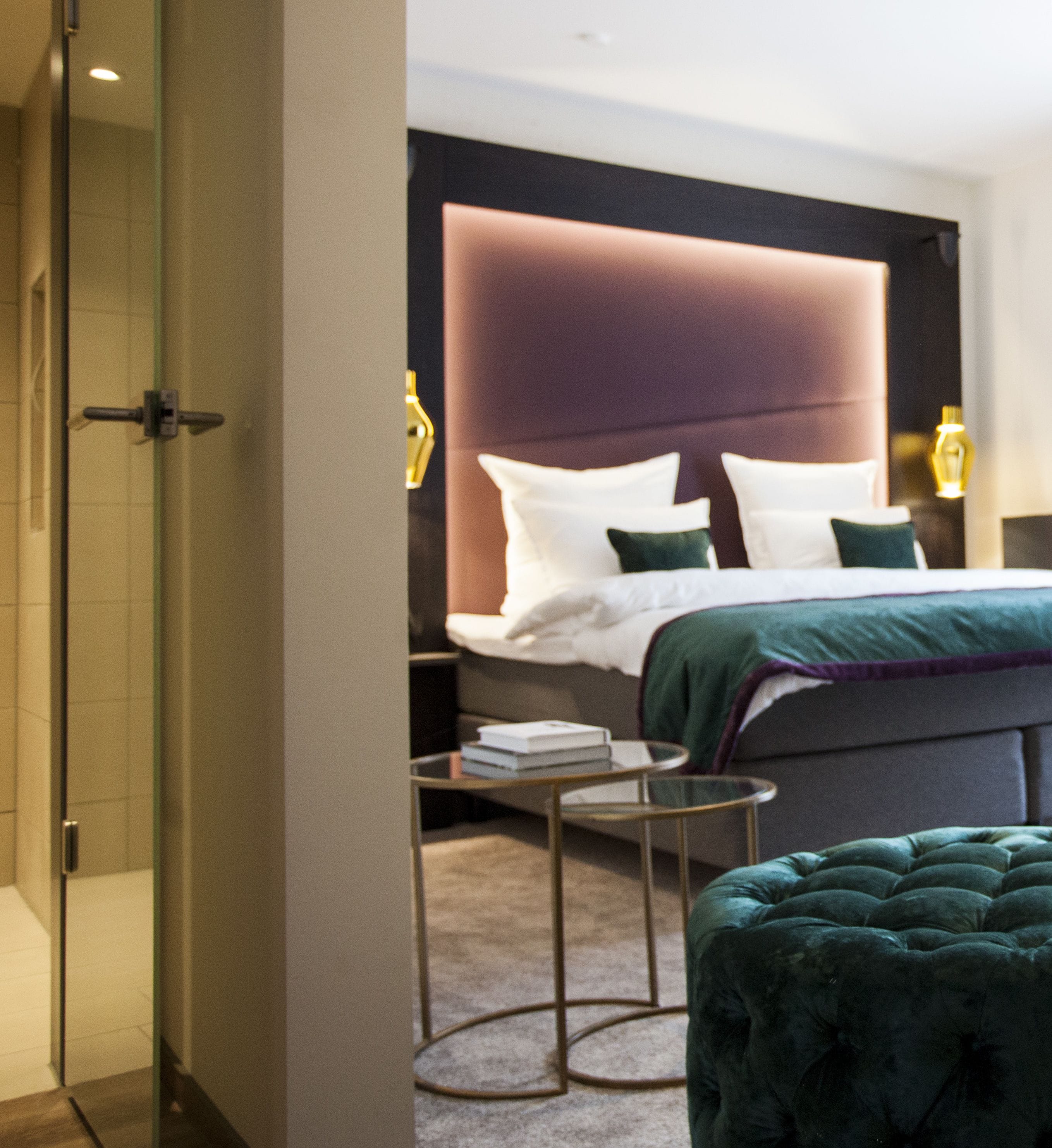 Una habitación de hotel moderna con una cama grande, una mesita de noche, una lámpara de lectura y un banco tapizado en verde.