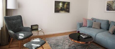 Een woonkamer met een blauwe bank, een grijze fauteuil, een klein bijzettafeltje, een lamp en een ingelijste foto aan de muur.