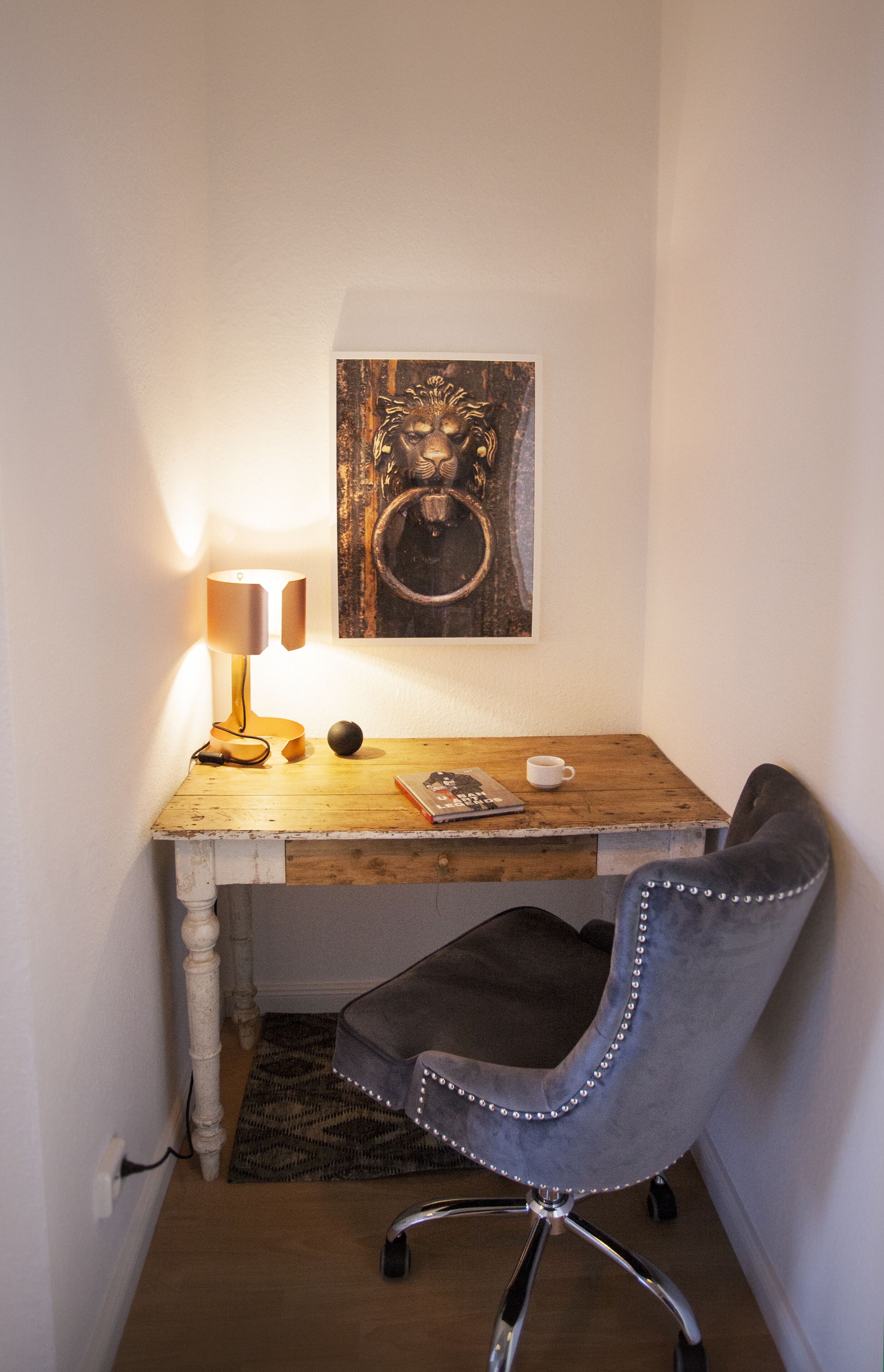 Un angolo studio accogliente con una scrivania in legno, una lampada moderna, una sedia grigia, un tappeto piccolo e un quadro appeso alla parete.