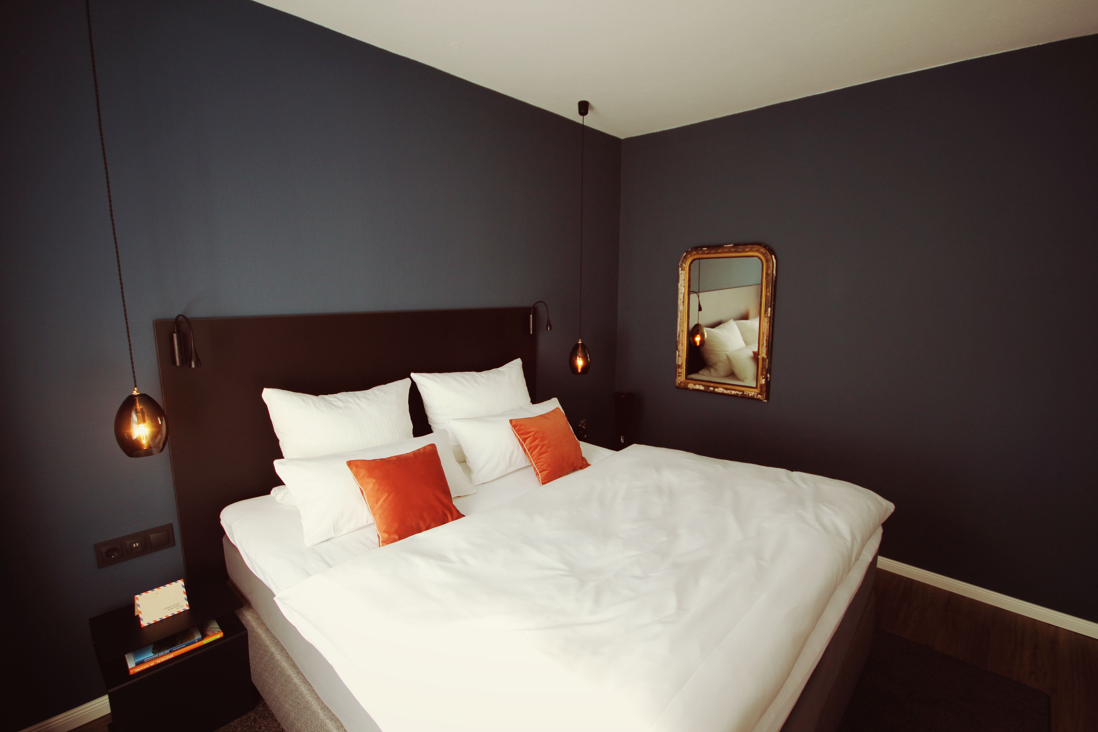 A neatly made bed with white and orange pillows, a dark headboard, and a bedside table with a book and a lamp.