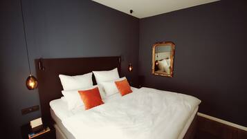 A neatly made bed with white and orange pillows, a dark headboard, and a bedside table with a book and a lamp.