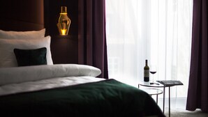 Una habitación de hotel con cama, mesita de noche con una botella de vino y una copa, y una ventana con cortinas.